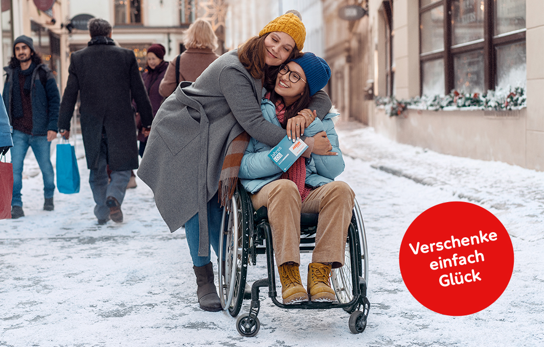 Eine Mutter und ihre Tochter, die im Rollstuhl sitzt, stehen mitten in der verschneiten Stadt und umarmen sich. Das Mädchen hält ein Jahreslos in der Hand.