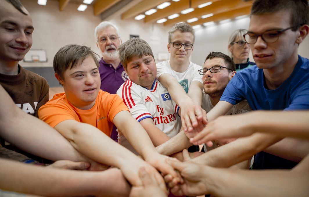 Eine Gruppe von Sportler*innen steht im Kreis und legt in der Mitte ihre Hände zusammen. Manche Personen haben eine Behinderung.