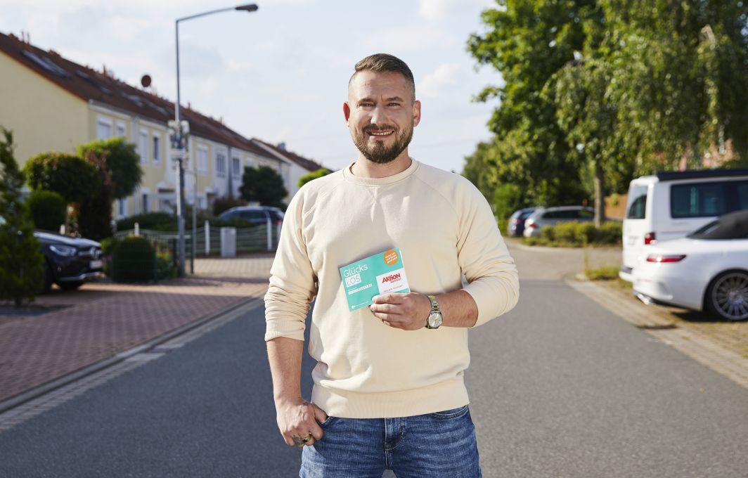 Ein Mann steht auf einer Straße und hält ein Glück-Los vor dem Bauch.