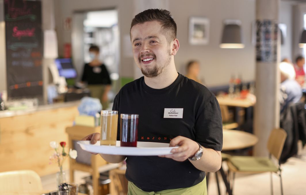 Ein junger Mann trägt ein Tablett mit zwei Gläsern. Er lacht.