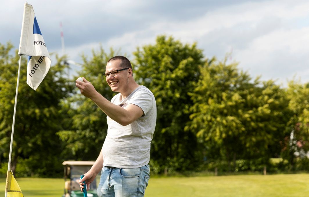 Ein junger Mann steht an einer Fahne auf dem Golfplatz und hält lachend einen Golfball hoch.