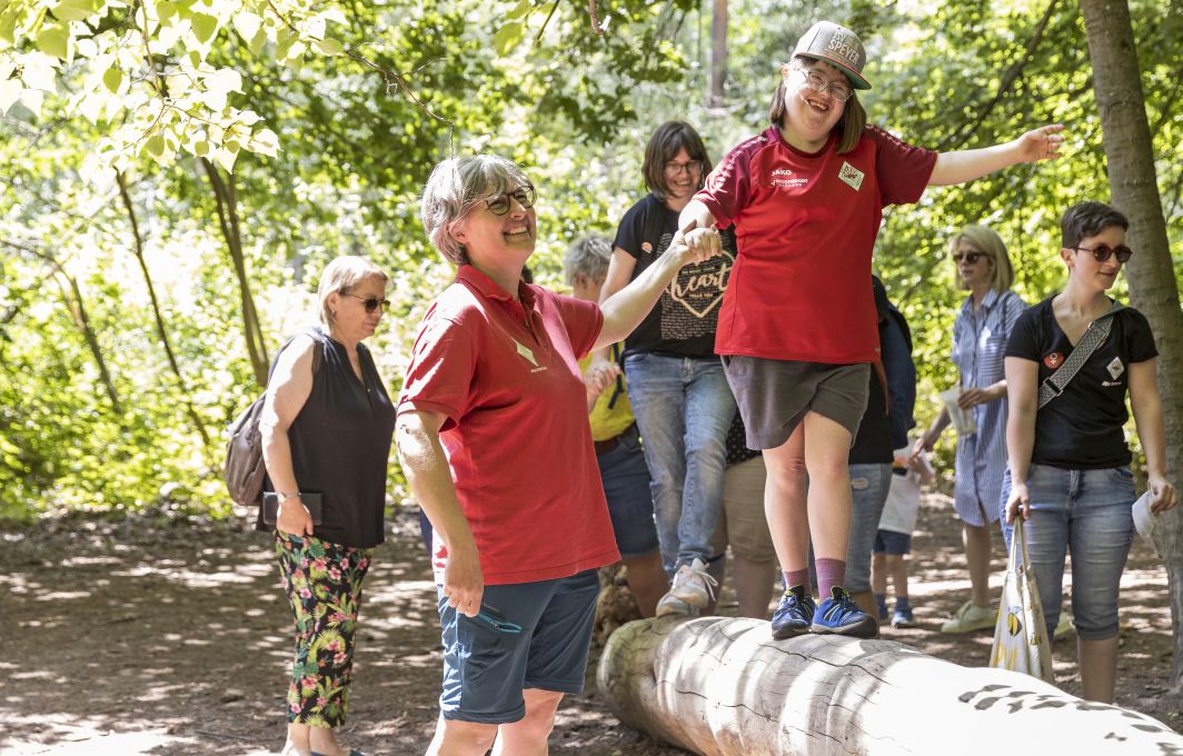 Ein Mädchen mit Down-Syndrom balanciert über einen Baumstamm im Wald. Eine Frau hilft ihr und hält sie an der Hand.