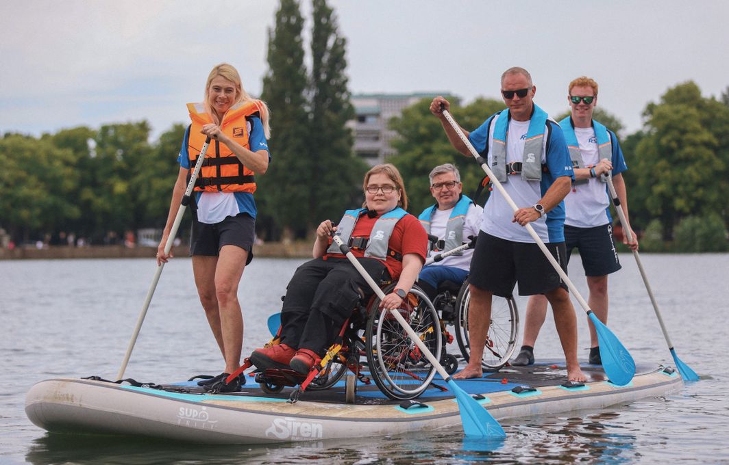 Fünf Personen sind auf einem Paddling-Board im Wasser. Zwei sitzen im Rollstuhl.