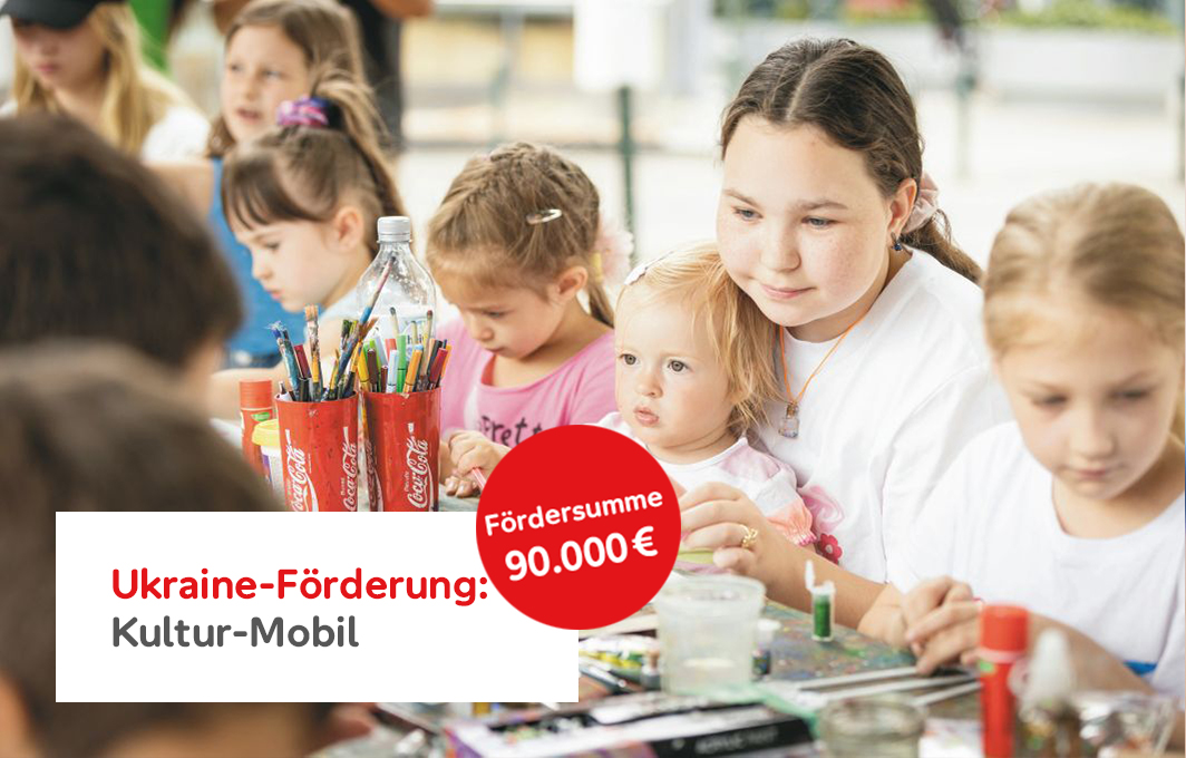 Mehrere Kinder sitzen am Tisch und haben Mal- und Bastelutensilien vor sich. Im Fokus ist ein älteres Mädchen mit einen kleineren Kind auf dem Schoß.