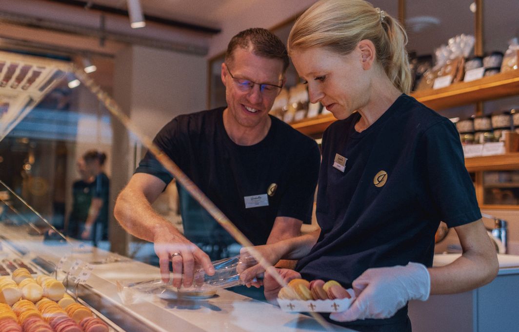 In einer Patisserie verpacken ein Mann und eine Frau eine Schachtel bunter Macarons.