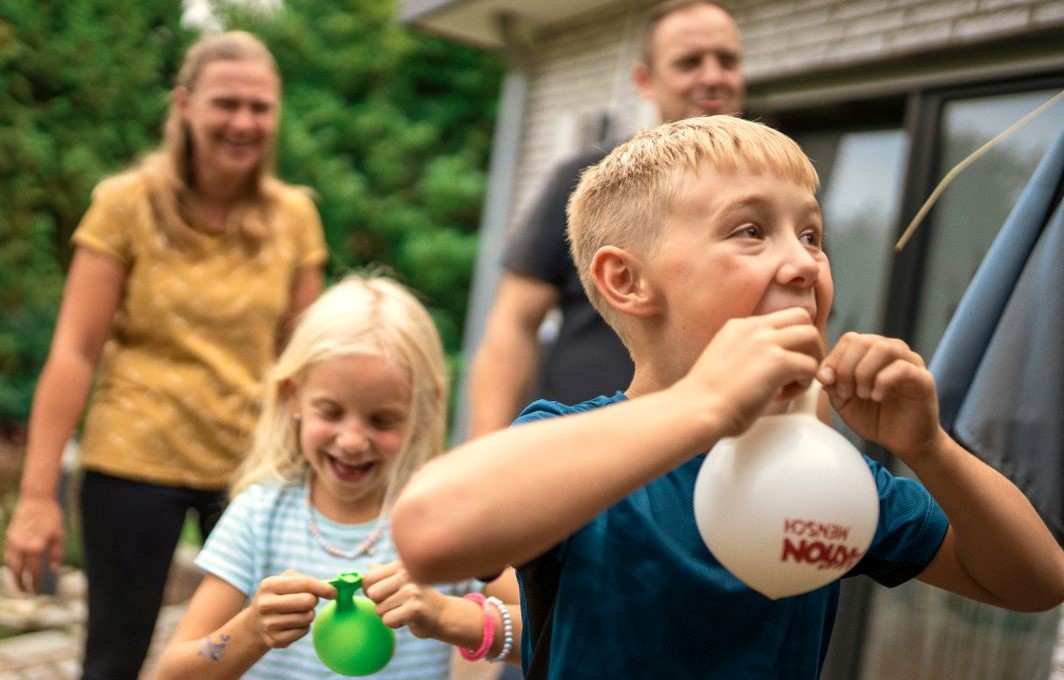 Zwei Kinder spielen lachend mit Luftballons, im Hintergrund sieht man unscharf ihre fröhlich lächelnden Eltern.