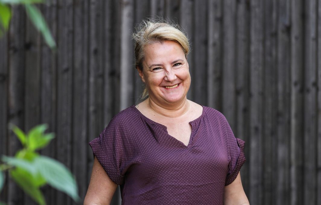 Eine Frau mit blondem Zopf und gepunktetem Shirt grinst fröhlich in die Kamera.