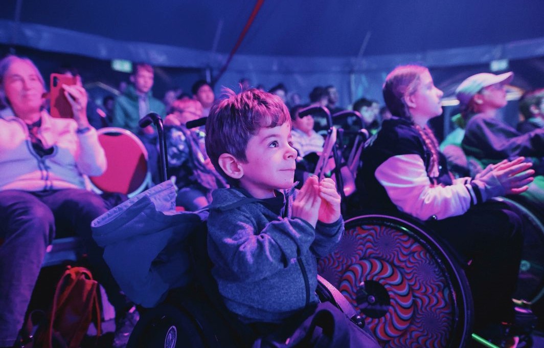 Ein Junge sitzt bei atmosphärischem Licht im Zirkus und klatscht Beifall. Er und weitere Kinder um ihn herum sitzen im Rollstuhl.