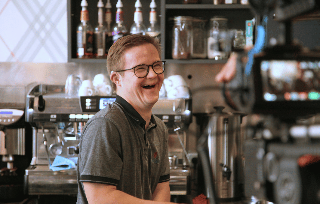 Ein junger Mann mit Downsyndrom steht in einem Cafe hinter der Theke und lacht.