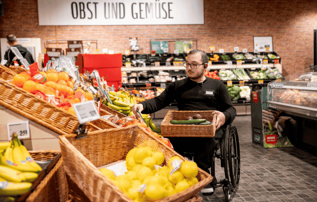 Ein Mann sitzt in einem Supermarkt im Rollstuhl und sortiert Obst und Gemüse ein.