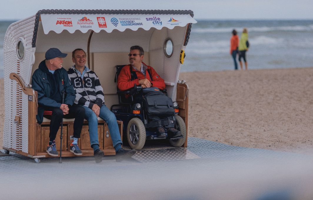 Drei Männer sitzen in einem Strandkorb am Meer. Einer von sitzt mit seinem Rollstuhl im Strandkorb, davor ist eine Rampe zur Auffahrt sichtbar.