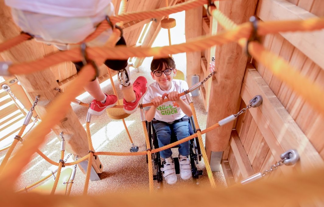 Ein Junge mit Brille und Rollstuhl auf dem inklusiven Spielplatz von oben fotografiert. Im linken Bildausschnitt ist die Beinprothese eines weiteren Kindes zu sehen, das auf einem Kletterseil steht.