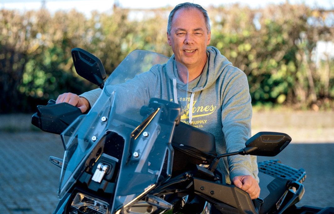 Ein Mann in grauem Pullover stützt sich auf den Lenker seines Motorrads.