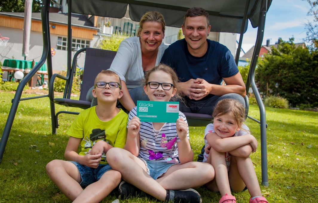 Familie Buhrmester sitzt in einer Hollywood-Schaukel in ihrem Garten. Die Eltern und die drei Kinder lachen in die Kamera und halten ein Glücks-Los in die Höhe.