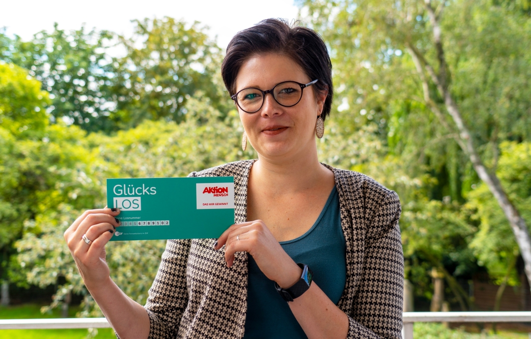 Eine Frau mit kurzen dunklen Haaren und Brille schaut lächelnd in die Kamera. Sie hält ein Glücks-Los in der Hand.