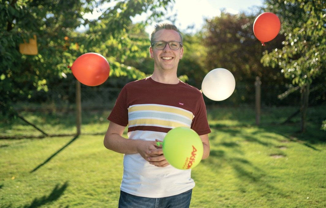 Ein junger Mann steht bei sonnigem Wetter im Garten. Um ihn herum fliegen bunte Luftballons.