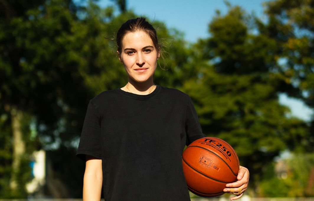 Eine junge Frau in schwarzer kurzer Sportkleidung. Sie hält einen Basketball unter dem Arm und lächelt in die Kamera.