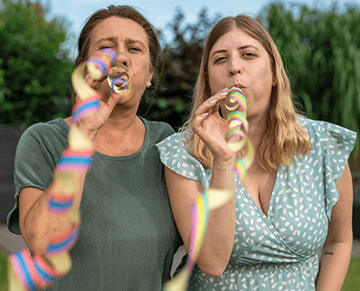 Zwei Frauen stehen nebeneinander in einem Garten. Sie schauen in die Kamera und pusten bunte Luftschlangen in die Luft.