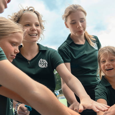 Die jugendlichen Spielerinnen einer inklusiven Hockey-Mannschaft stehen im Kreis auf einem Sportplatz. Sie haben ihre Hände in die Mitte gestreckt und lachen fröhlich.