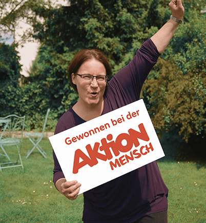 Eine Frau hält ein Schild in ihrer einen Hand, auf dem steht 'Gewonnen bei der Aktion Mensch', ihren anderen Arm streckt sie jubelnd in die Luft.