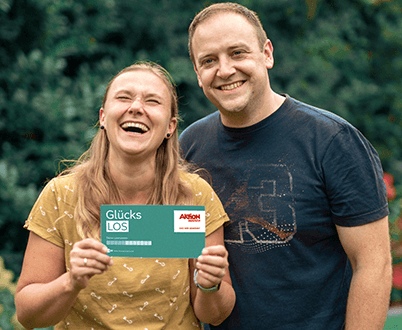 Die Gewinner*innen Bozena und Sebastian stehen in sommerlicher Kleidung nebeneinander in ihrem Garten. Bozena hält ihr Glücks-Los in die Kamera und lacht.