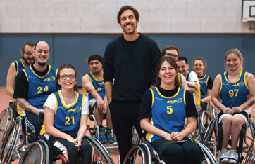 In einer Sporthalle steht Max Giesinger inmitten einer Gruppe von Rollstuhl-Basketballer*innen. Alle schauen freundlich in die Kamera und lächeln.