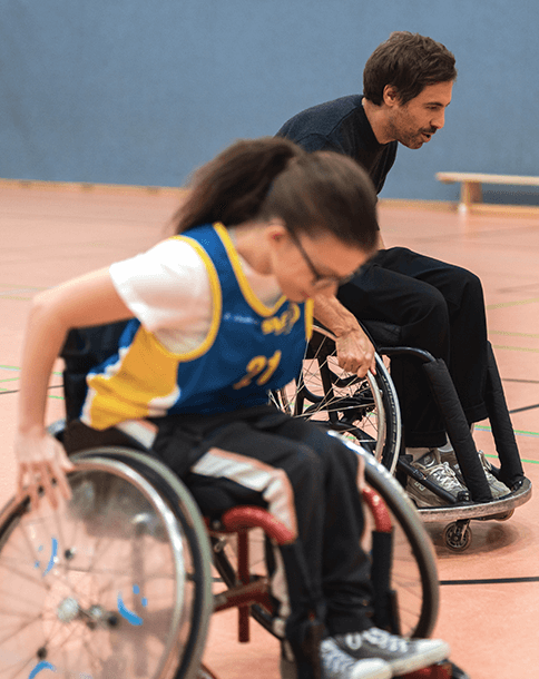 In einer Sporthalle sieht man den Sänger Max Giesinger gemeinsam mit einer jungen Rollstuhlbasketball-Spielerin. Beide sitzen in einem Sportrollstuhl und treten in einem Wettfahren gegeneinander an.