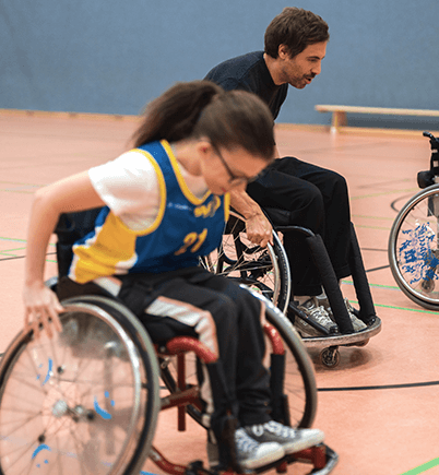 In einer Sporthalle sieht man den Sänger Max Giesinger gemeinsam mit einer jungen Rollstuhlbasketball-Spielerin. Beide sitzen in einem Sportrollstuhl und treten in einem Wettfahren gegeneinander an.