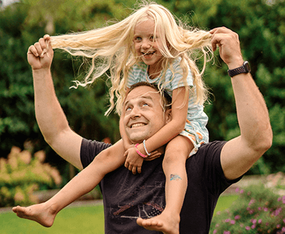 Die Gewinner*innen Bozena und Sebastian stehen in sommerlicher Kleidung nebeneinander in ihrem Garten. Bozena hält ihr Glücks-Los in die Kamera und lacht.