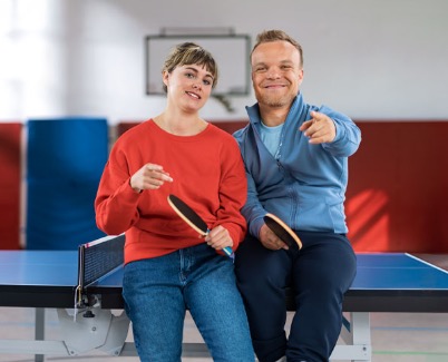 Niko Kappel und eine junge Frau sitzen entspannt auf einer Tischtennisplatte in einer Sporthalle. Beide halten Tischtennisschläger in der Hand und lächeln in die Kamera. Die Person links trägt einen roten Pullover und Jeans, Niko Kappel trägt eine blaue Sportjacke und zeigt mit einem Finger nach vorne.
